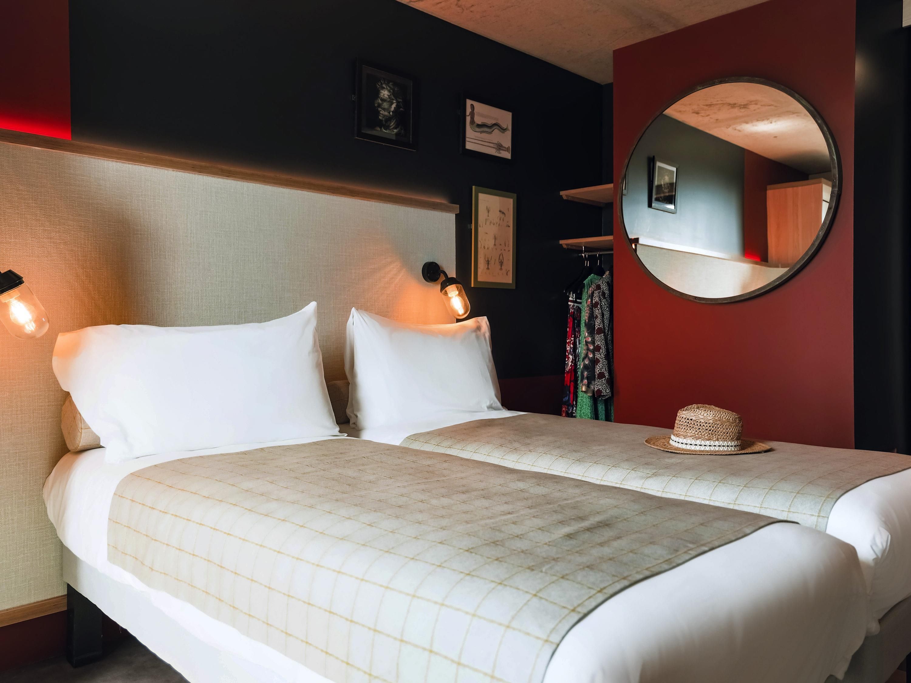 A hotel room with two beds, a round mirror, and a wall-mounted light fixture.