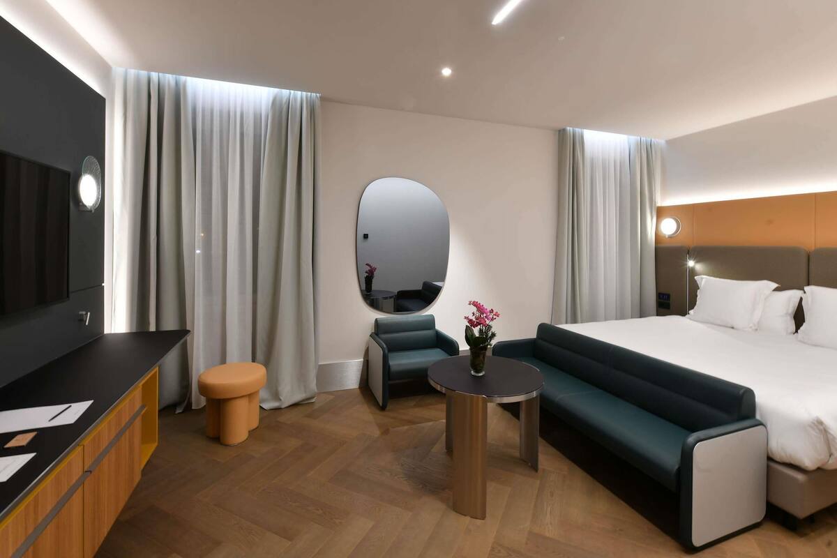 A modern hotel room with a bed, a round mirror, a small table with a vase, a sofa, and a TV.