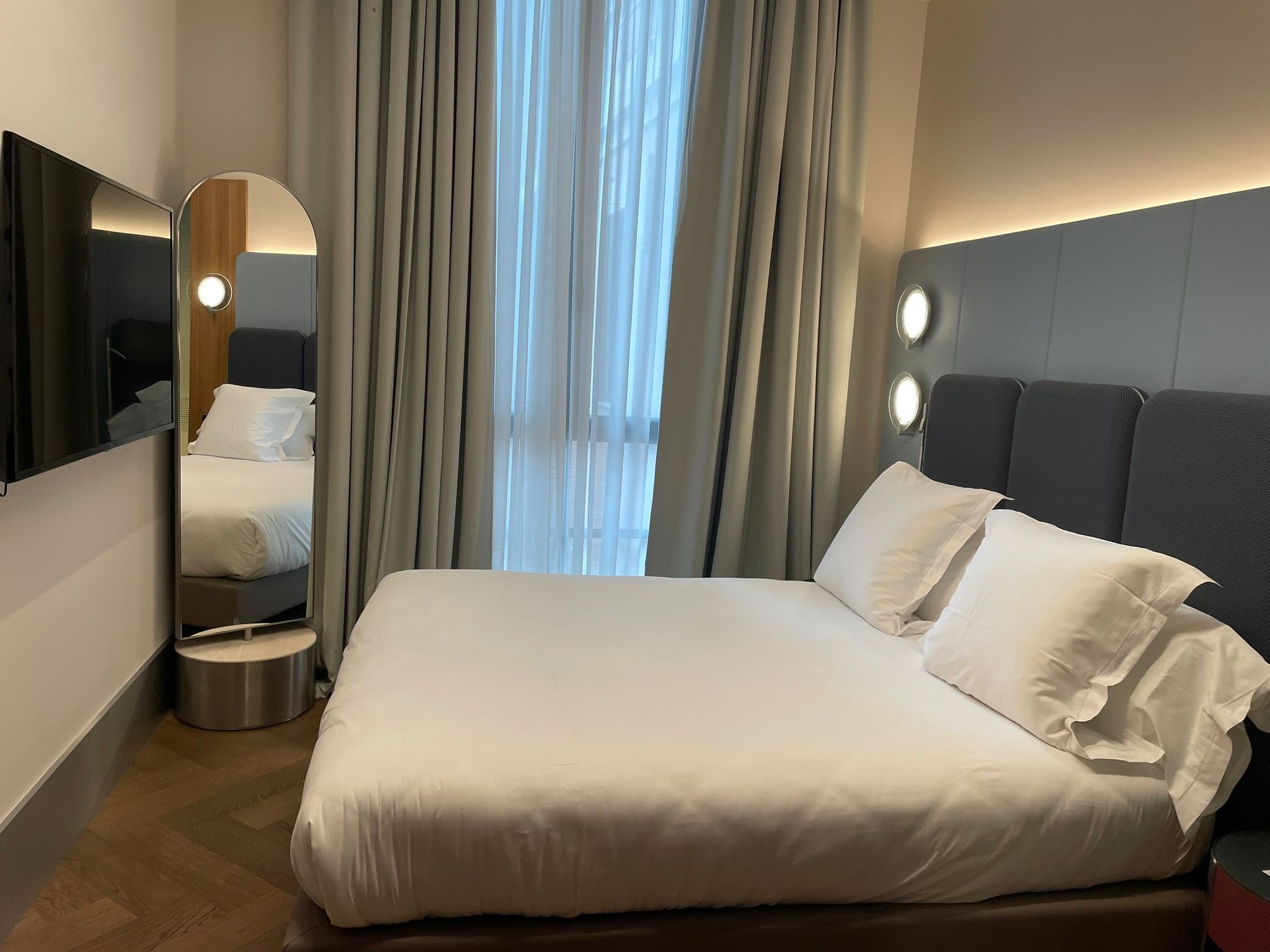 Una habitación de hotel moderna con una cama grande, un espejo redondo y vistas de la ciudad a través de cortinas translúcidas.
