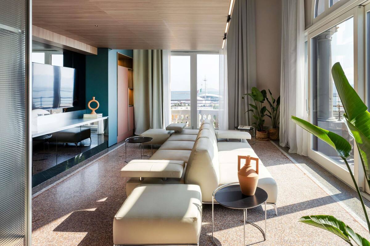 A modern living room with a long white sofa, a TV, and a large window with a city view.