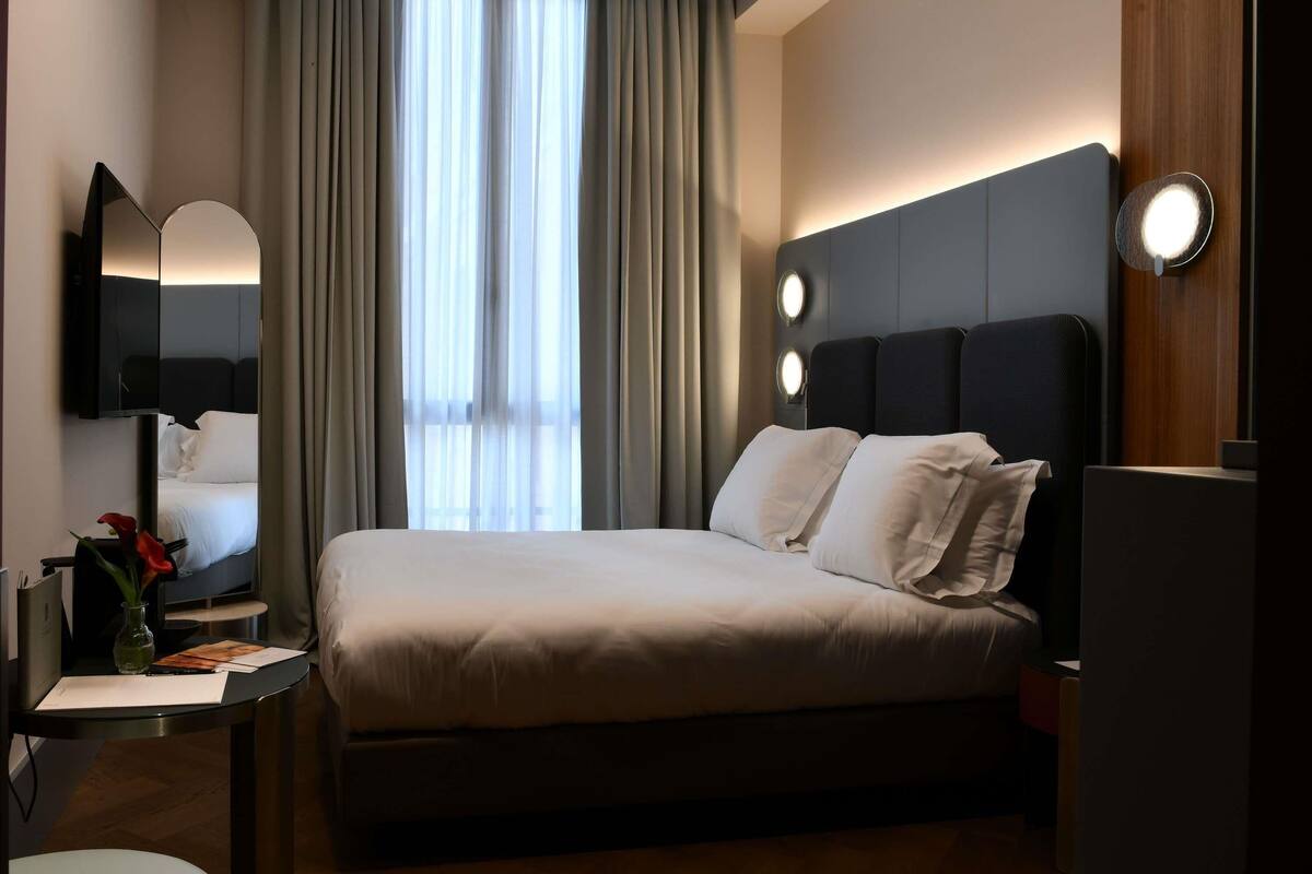 A modern hotel room with a large bed, a desk with a lamp, a mirror, and a window with curtains.