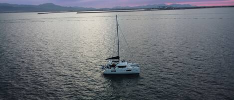 A sailboat named "PALAZZOTIRSO" on calm waters with distant mountains and industrial structures.