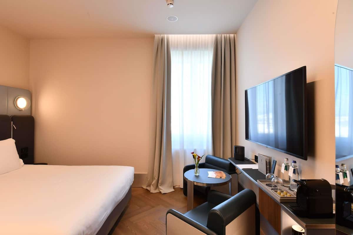 A hotel room with a bed, a desk with a flat-screen TV, a chair, a small table with a vase, and a window with curtains.