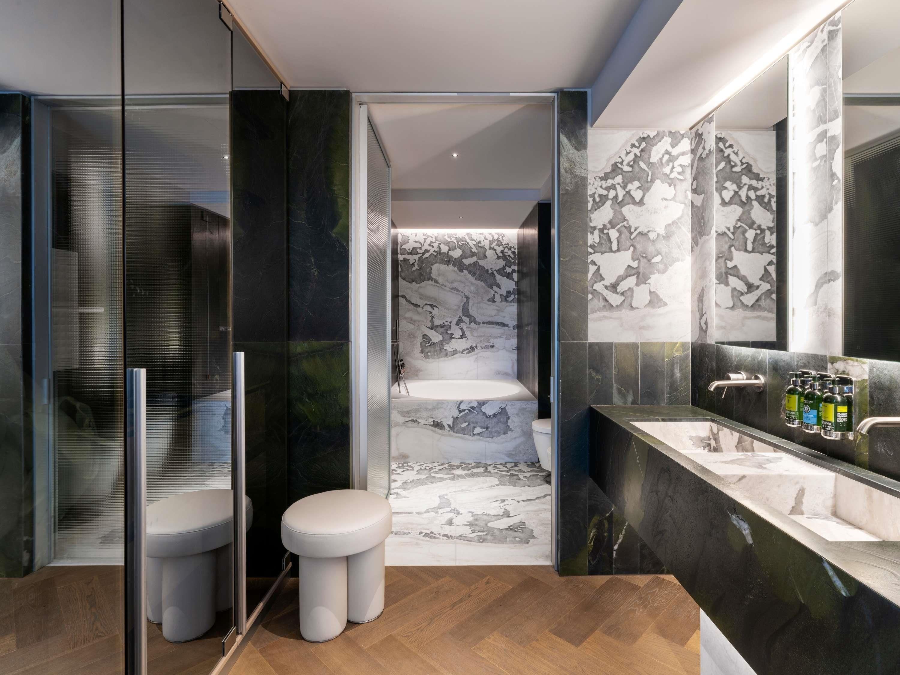 Een moderne badkamer met een douchecabine van glas, een wit toilet, een doucheruimte betegeld met marmer en een wastafel met een grote spiegel en toiletartikelen.