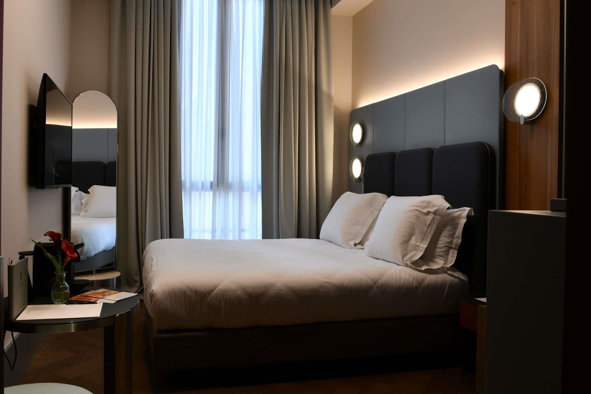 Une chambre d’hôtel moderne avec un grand lit, un bureau avec une lampe, un miroir et une fenêtre avec des rideaux.