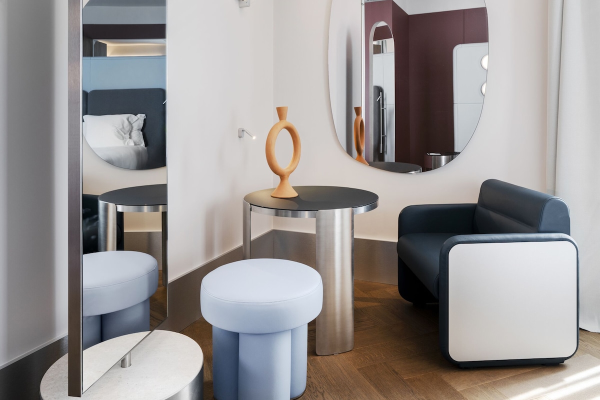 Een moderne hotelkamer met een grote spiegel, een ronde tafel, een blauwe fauteuil en een klein krukje.