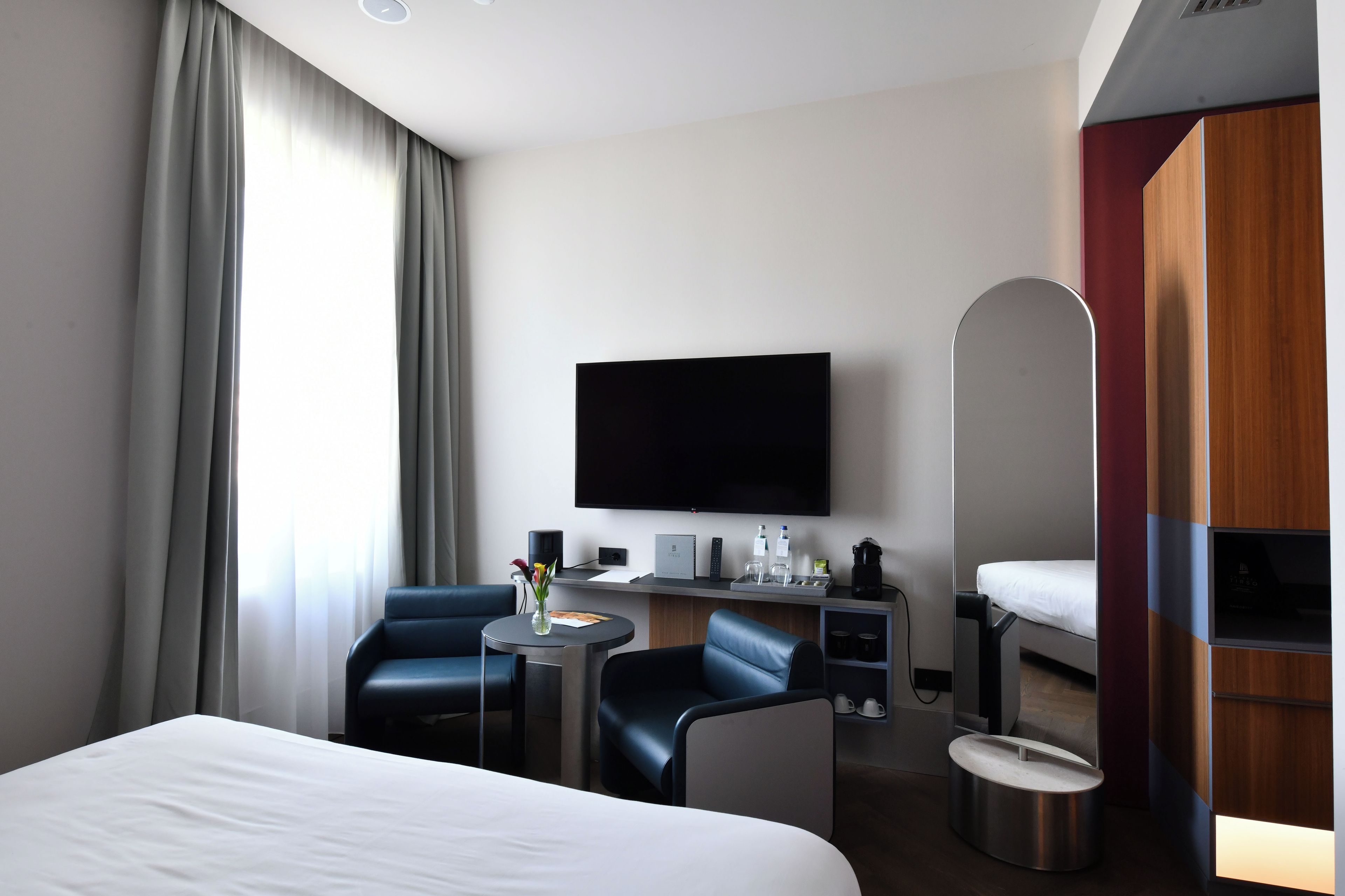 A hotel room with a bed, a desk with a flat-screen TV, a chair, a small table, and a minibar.