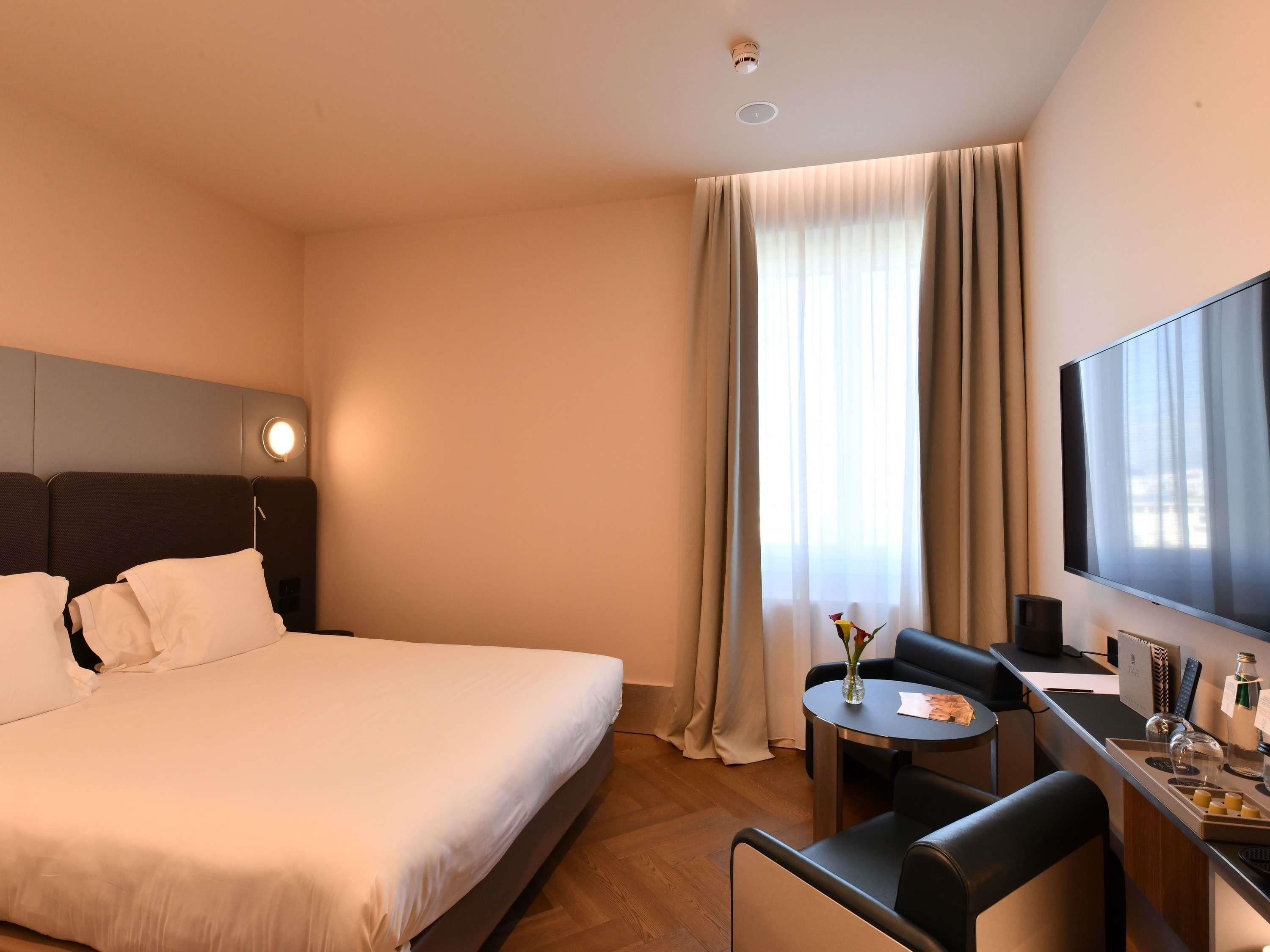 A hotel room with a bed, a desk with a chair, a TV, and a window with curtains.
