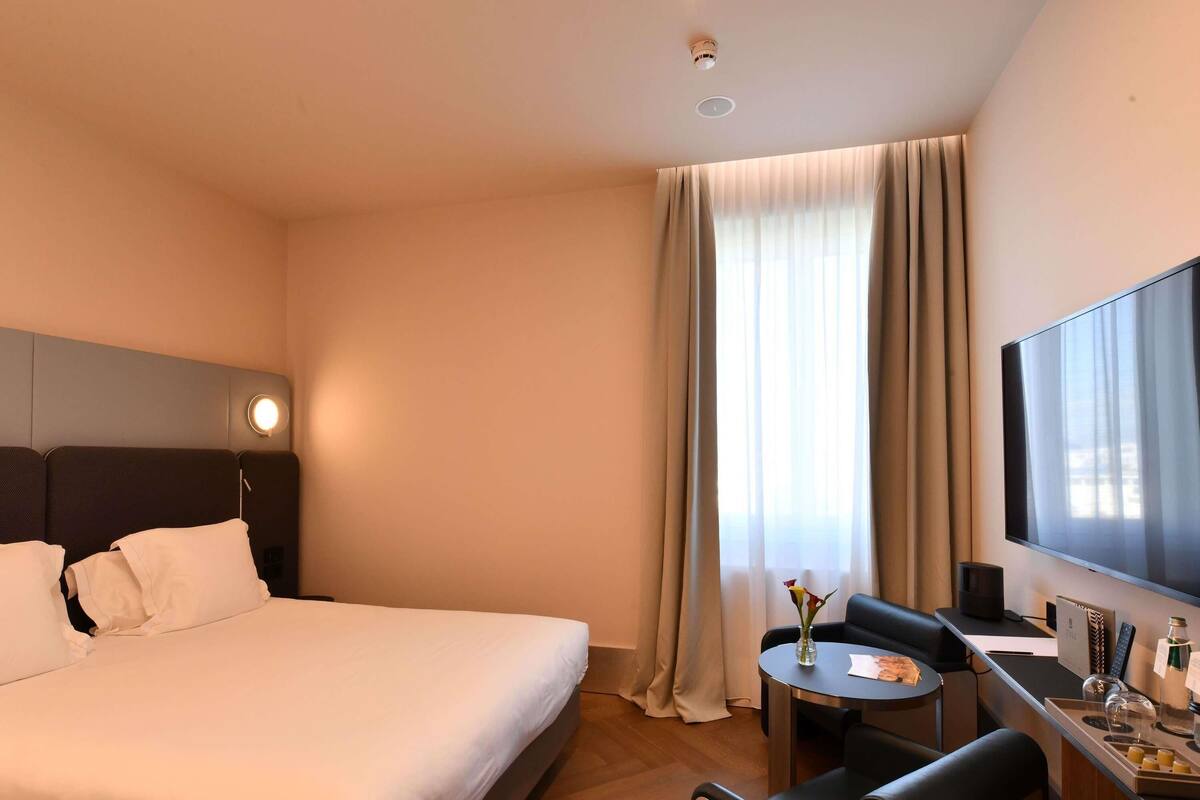 A hotel room with a bed, a desk with a chair, a TV, and a window with curtains.