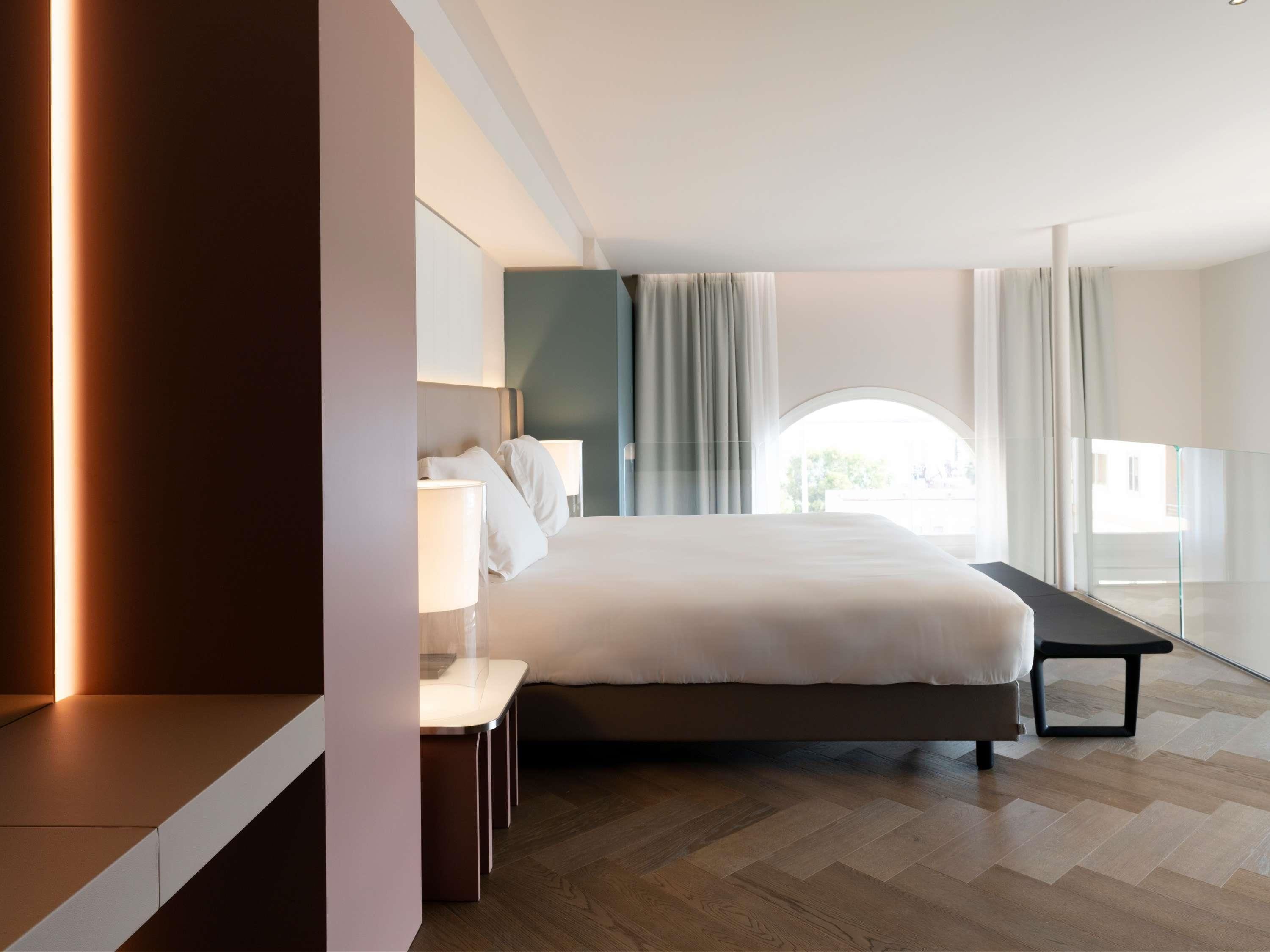 A modern hotel room with a large bed, bedside tables, a desk, and a view of the outdoors.