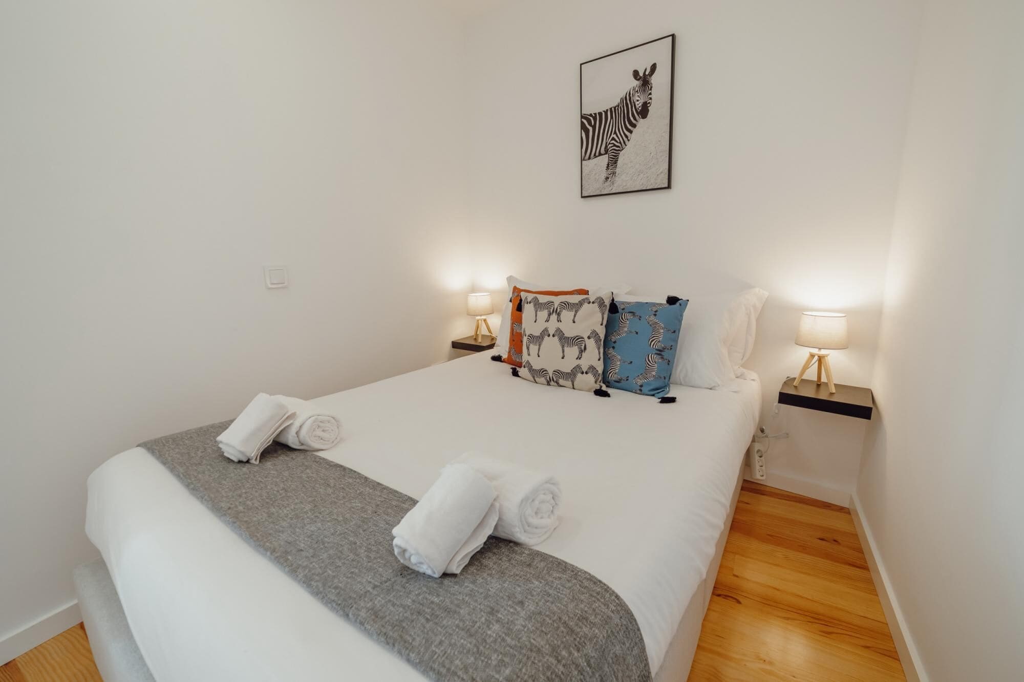 A neatly made bed with two rolled towels, two pillows with zebra prints, and two bedside lamps on wooden nightstands.