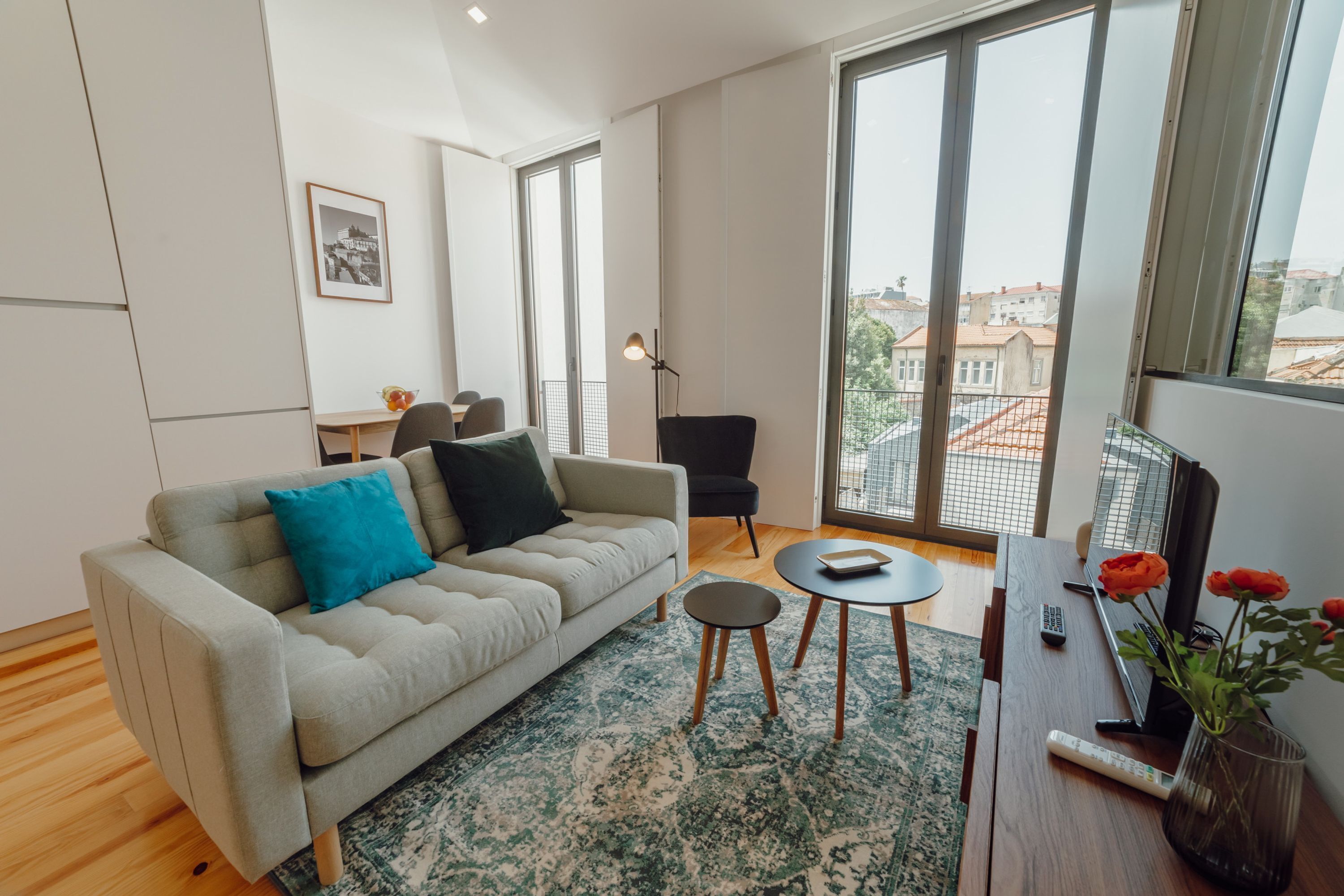 A modern living room with a sofa, a coffee table, and a TV. There is a large window with a view of buildings outside.