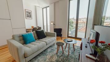 A modern living room with a sofa, a coffee table, and a TV. There is a large window with a view of buildings outside.