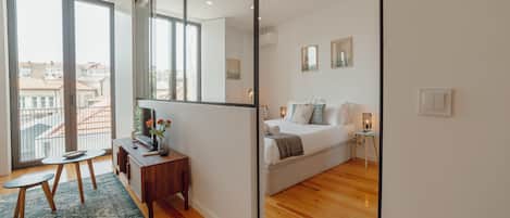 A modern bedroom with a large bed, a small desk, a chair, and a view of the city through large windows.
