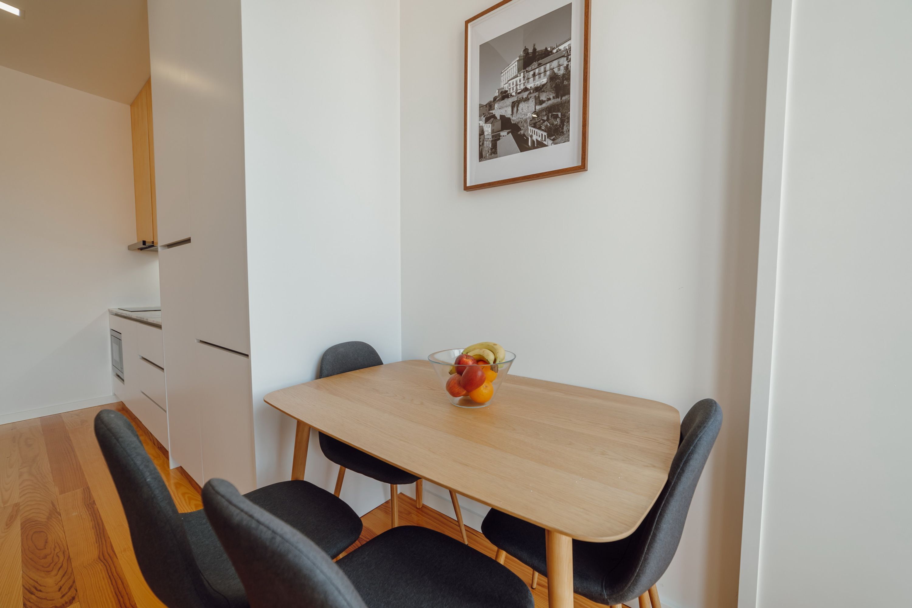 Een eetruimte met een houten tafel en vier stoelen, een fruitschaal op de tafel en een lijst met een afbeelding aan de muur.