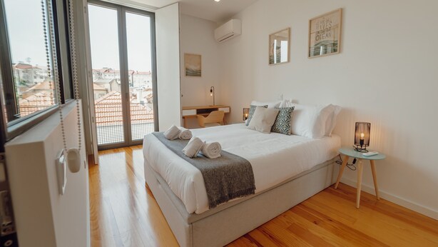 A bedroom with a large bed, a desk, a chair, and a view of the city through a large window. - Courtyard Oporto Design Apartment G (Porto)