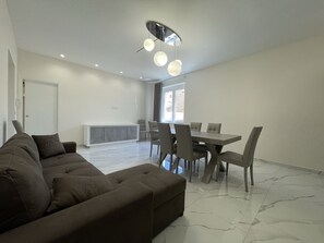 A modern living room with a sectional sofa, a dining table with chairs, and a ceiling light fixture.