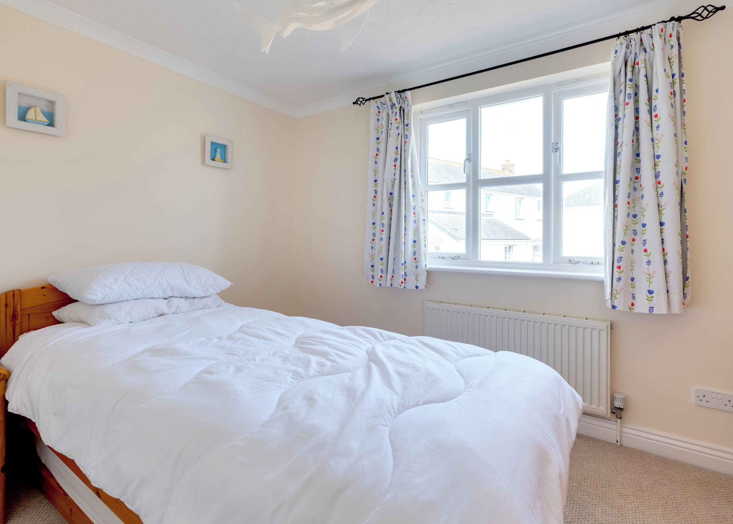 A bedroom with a bed, a window with floral curtains, a radiator, and two wall-mounted pictures.