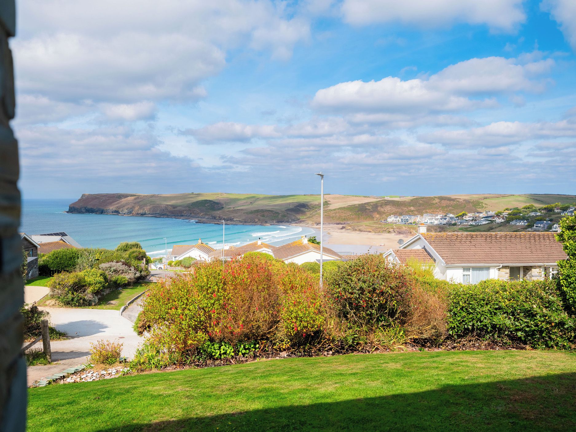 A coastal residential area with houses, a clear view of the sea, and a hilly landscape in the background.