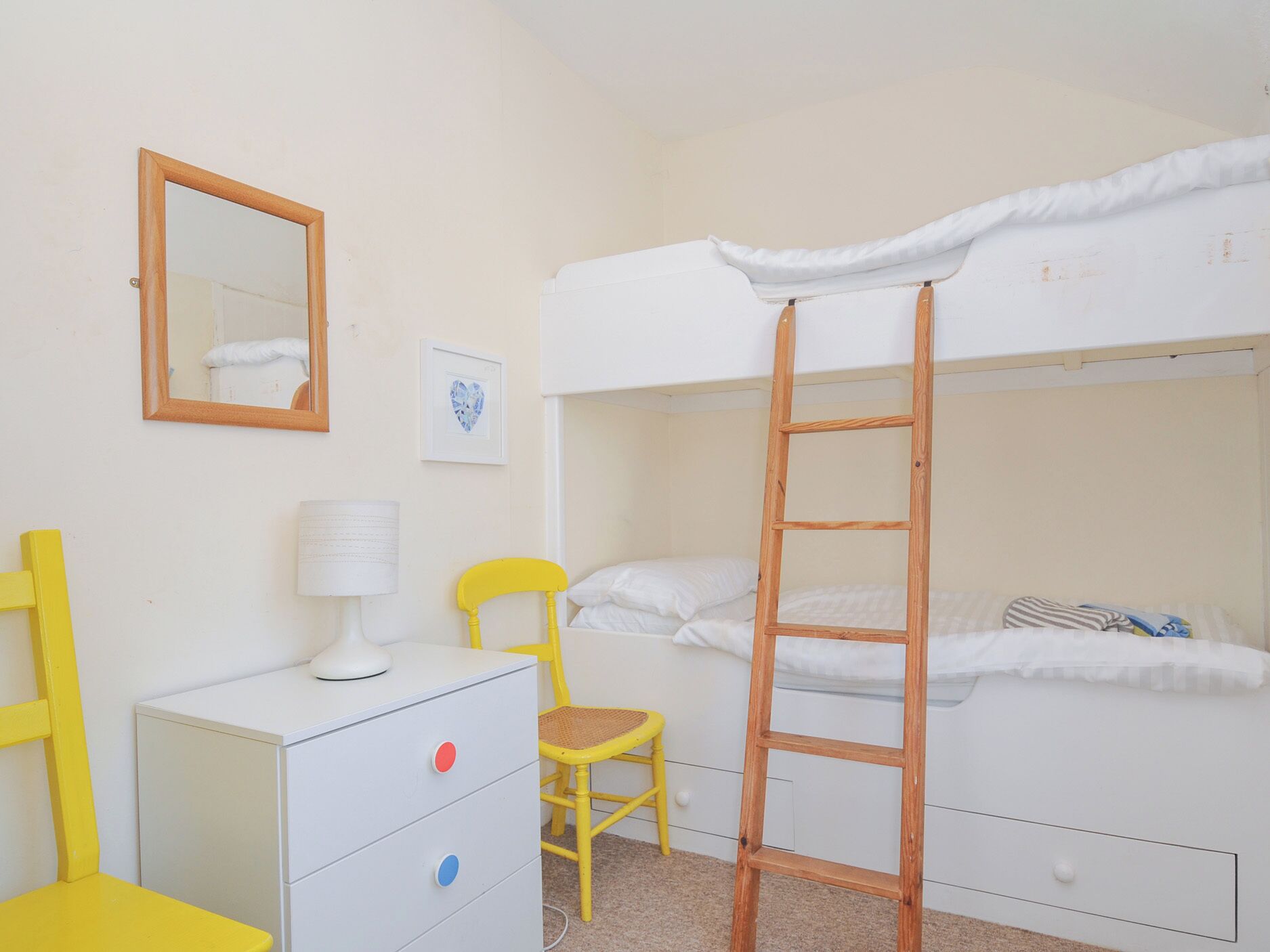 A bunk bed room with a ladder, a desk with drawers, a chair, a lamp, and a mirror on the wall.