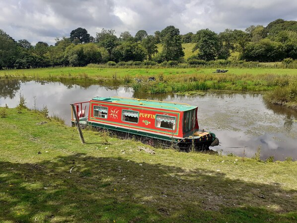 A stationary boat with 'PUFFIN' written on it, docked at a calm river with grassy banks and trees in the background. - Pretty & Cosy Boat in Stunning Valley View, Wales (Rhayader)