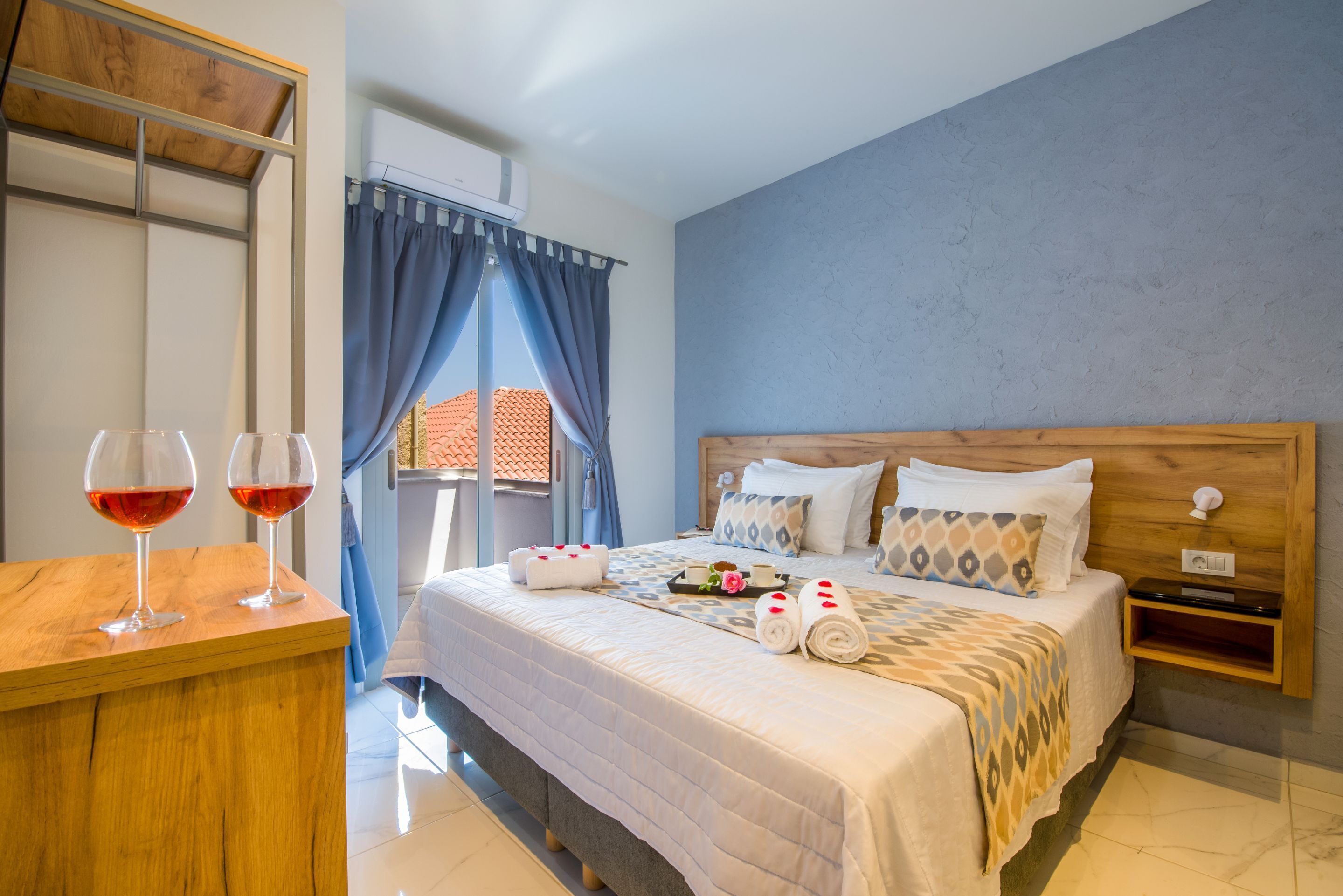 A hotel room with a bed, a wooden nightstand, a desk with two glasses of red wine, and a window with blue curtains.