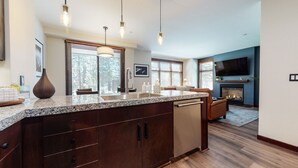 A modern kitchen with granite countertops, dark wood cabinets, and a built-in dishwasher. There are pendant lights above the island, and the kitchen opens into a living area with a fireplace and a flat-screen TV mounted on the wall. - Solstice #14 2 Bedroom Condo by RedAwning (Mammoth Lakes)