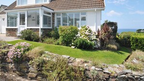 Une maison à deux étages, avec un toit en tuiles, des fondations en pierre et un jardin bien entretenu où poussent différentes plantes et fleurs.