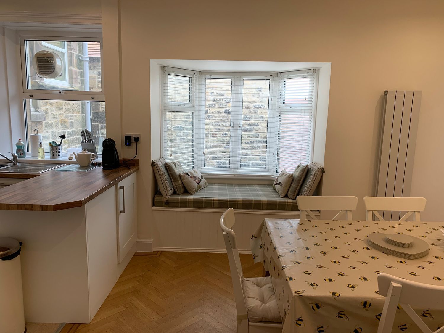 A cozy kitchen with a bay window seating area, a dining table with a bee-patterned tablecloth, and a radiator on the wall.