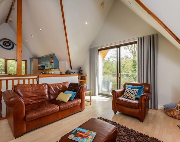 A spacious living area with a high ceiling, a wooden ceiling beam, a brown leather sofa, a wooden coffee table, and a dining area with a kitchen in the background.