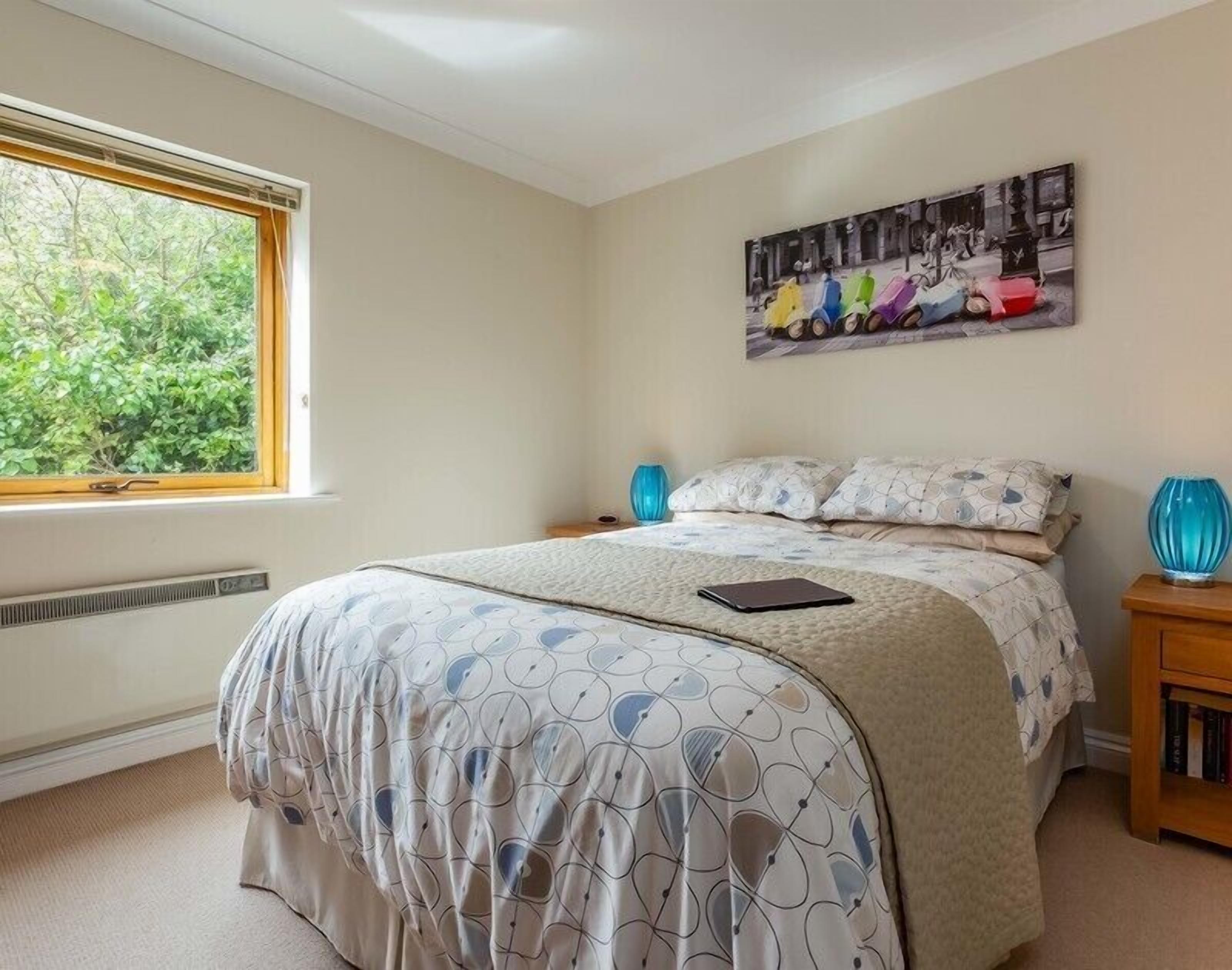 Een slaapkamer met een groot raam, een bed met beddengoed met een patroon, een nachtkastje met een lamp, en een aan de muur hangend schilderij.