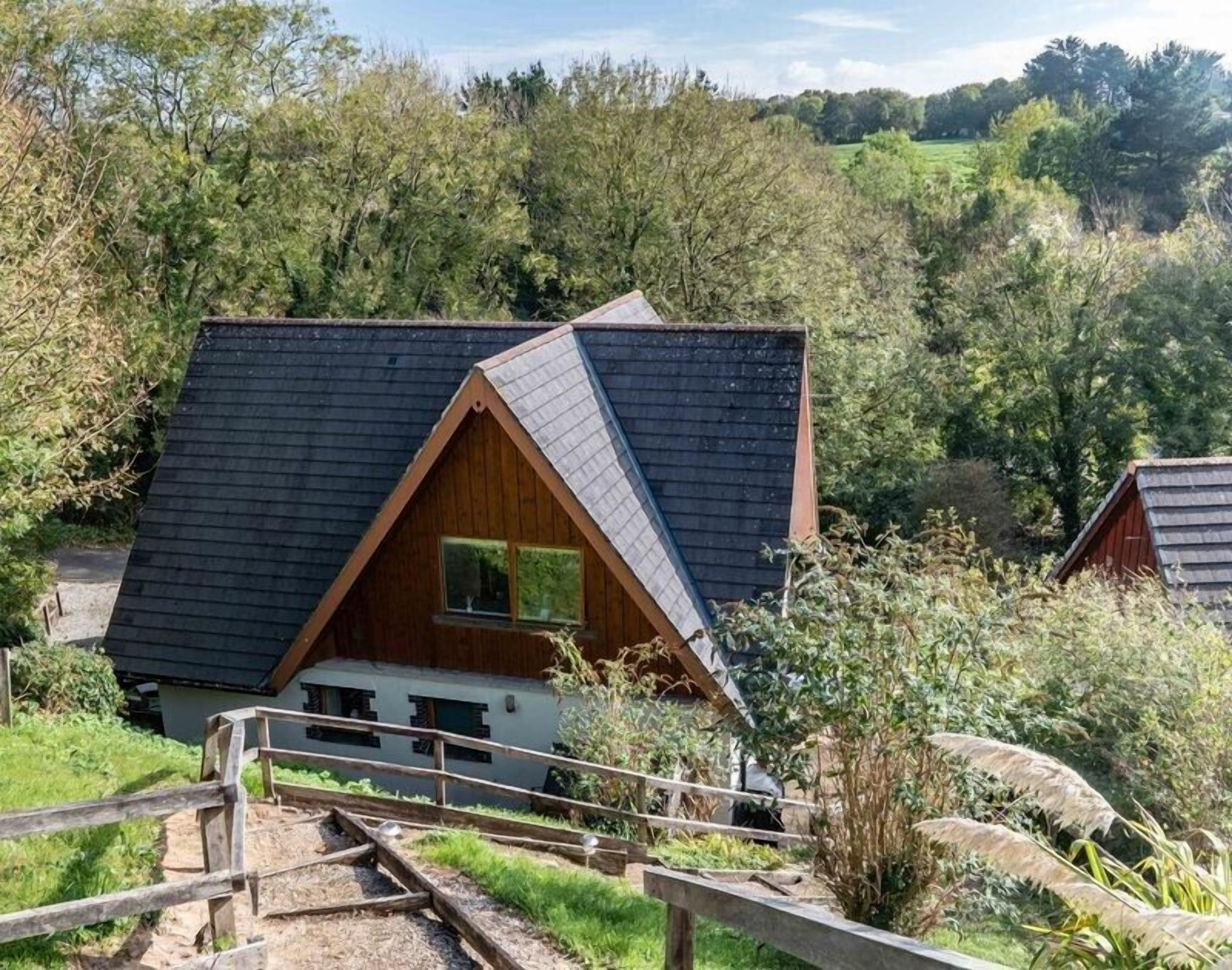 A small, triangular-roofed cabin with a wooden exterior, surrounded by lush greenery and a clear sky.