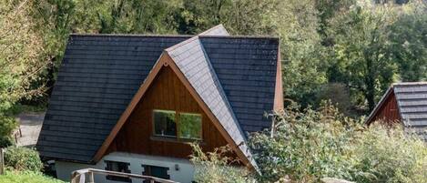 A small, triangular-roofed cabin with a wooden exterior, surrounded by lush greenery and a clear sky.