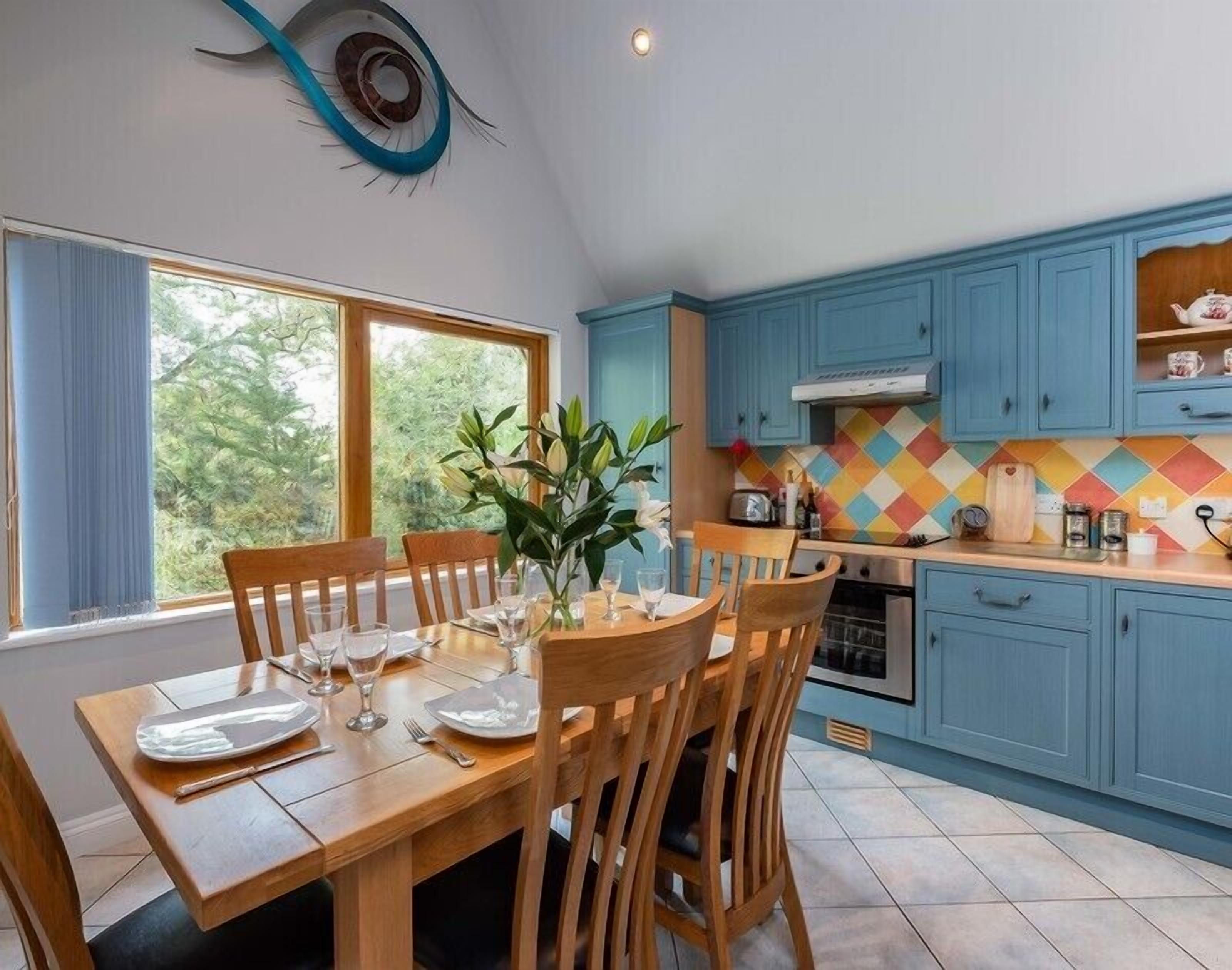 A dining area with a wooden table set for four, a window with curtains, and a kitchen with blue cabinets and a colorful backsplash.