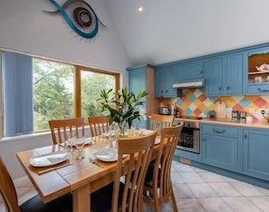 A dining area with a wooden table set for four, a window with curtains, and a kitchen with blue cabinets and a colorful backsplash.