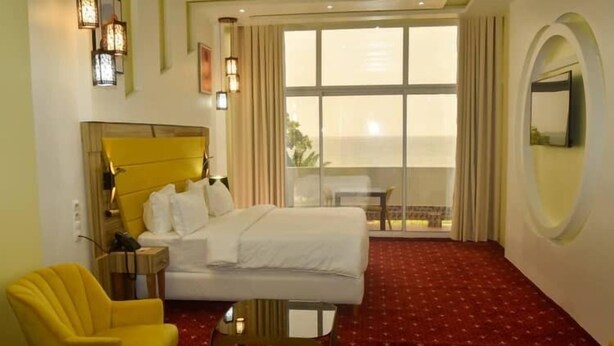 Quarto de hotel com uma cama, poltrona amarela, mesa pequena, janela grande com cortinas e espelho redondo na parede.