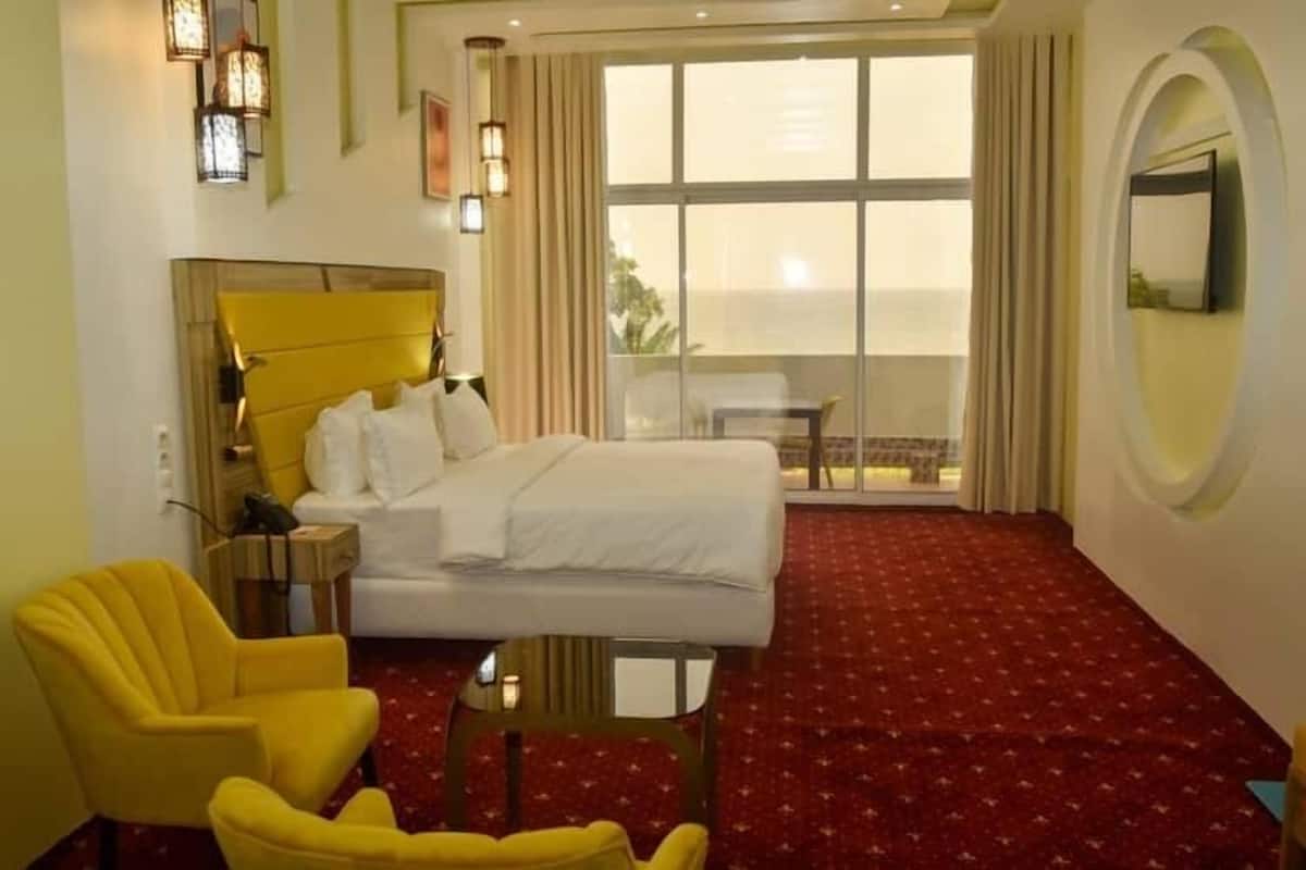 Een hotelkamer met een bed, een gele fauteuil, een klein tafeltje, een groot raam met gordijnen en een ronde spiegel aan de muur.