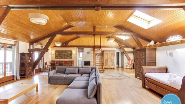 A spacious loft-style room with a wooden ceiling, a gray sectional sofa, a wooden bed, and a large window providing natural light.