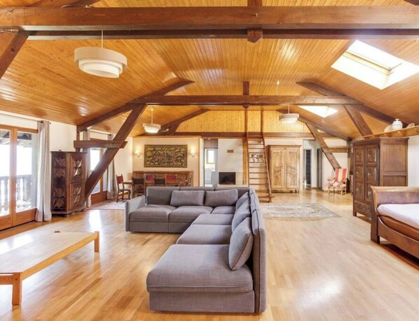 A spacious loft-style room with a wooden ceiling, a gray sectional sofa, a wooden bed, and a large window providing natural light. - Bergerie – Loft Apartment with Panoramic Lake & Mountain Views (Talloires-Montmin)