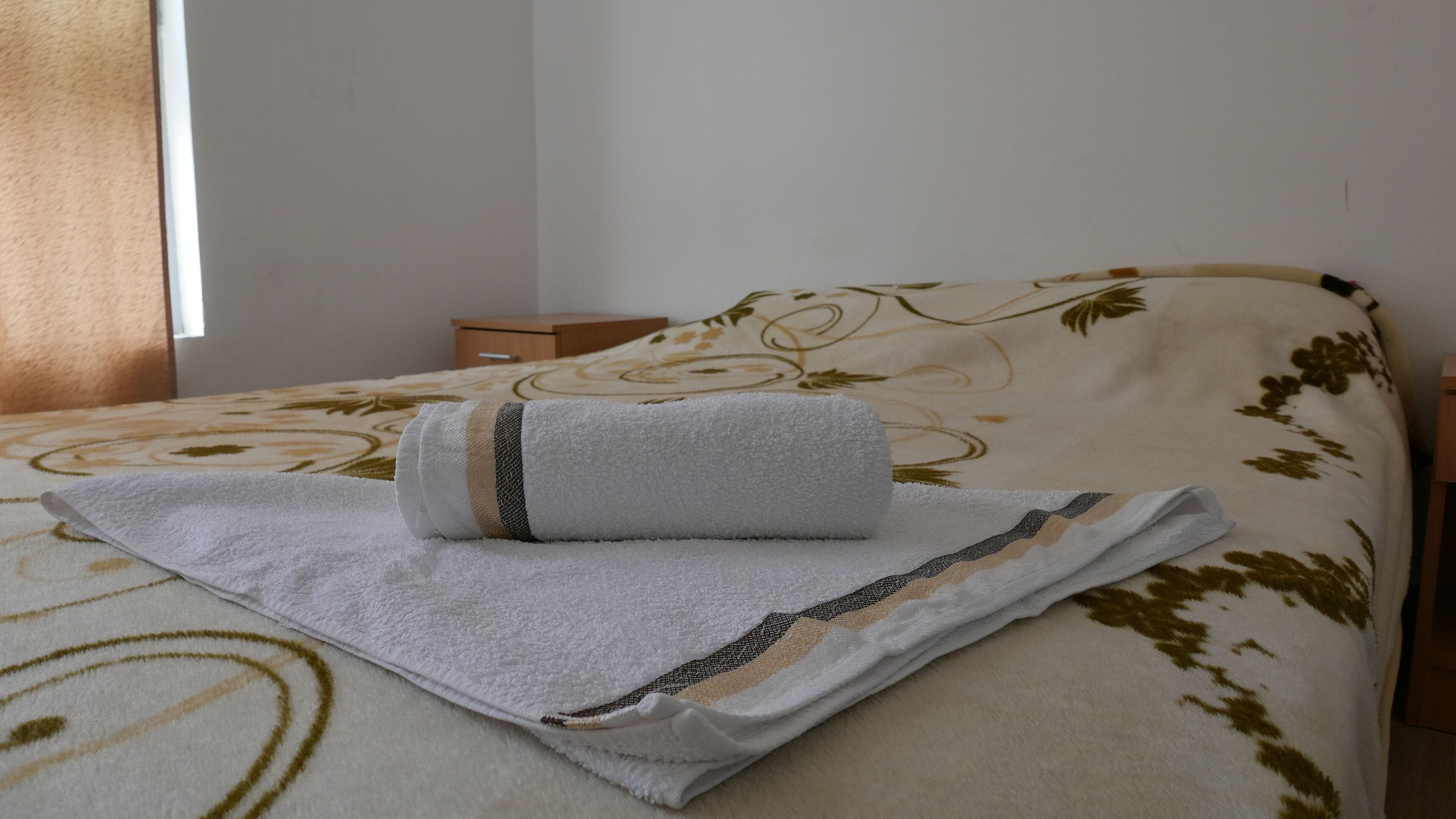 A neatly made bed with a white and beige floral bedspread, a rolled white towel, and a small wooden nightstand with a drawer.