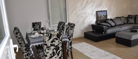 A modern living room with a dining area, featuring a sofa set, a dining table with chairs, and a large window with blinds.