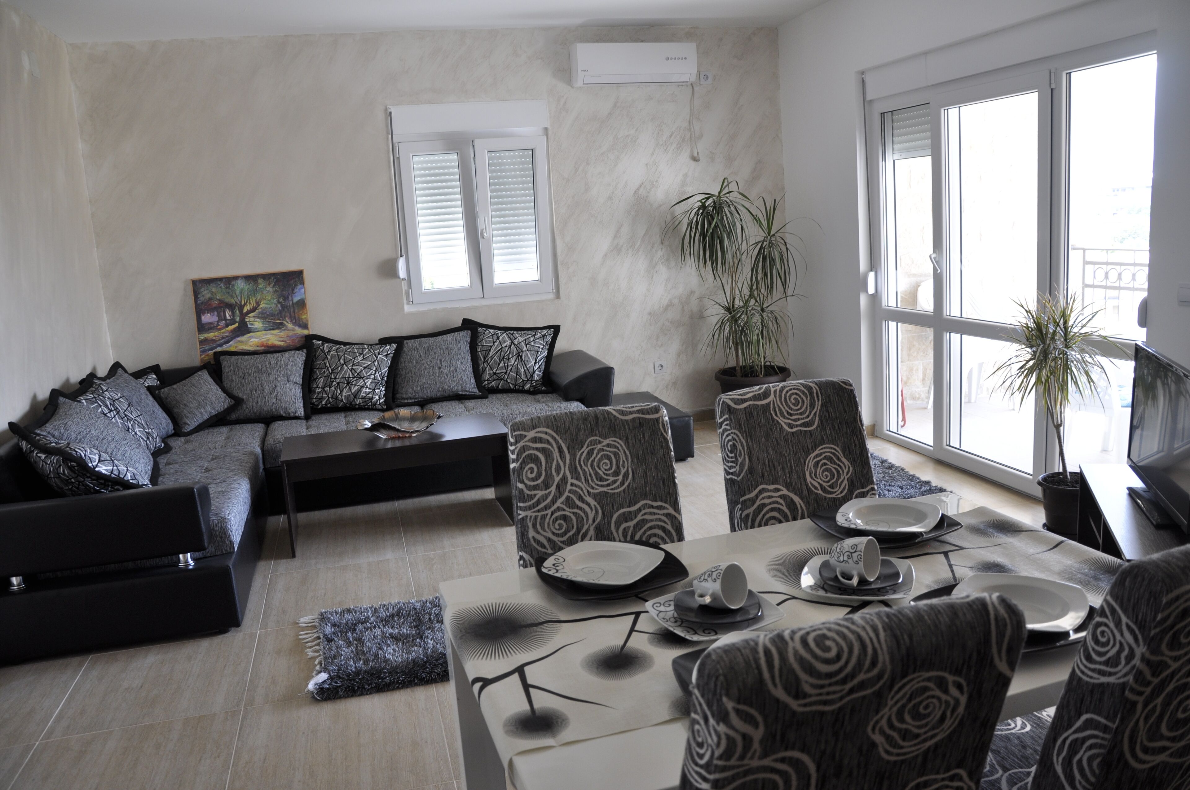 A modern living room with a sectional sofa, patterned armchairs, a dining table set for two, and large windows with blinds.