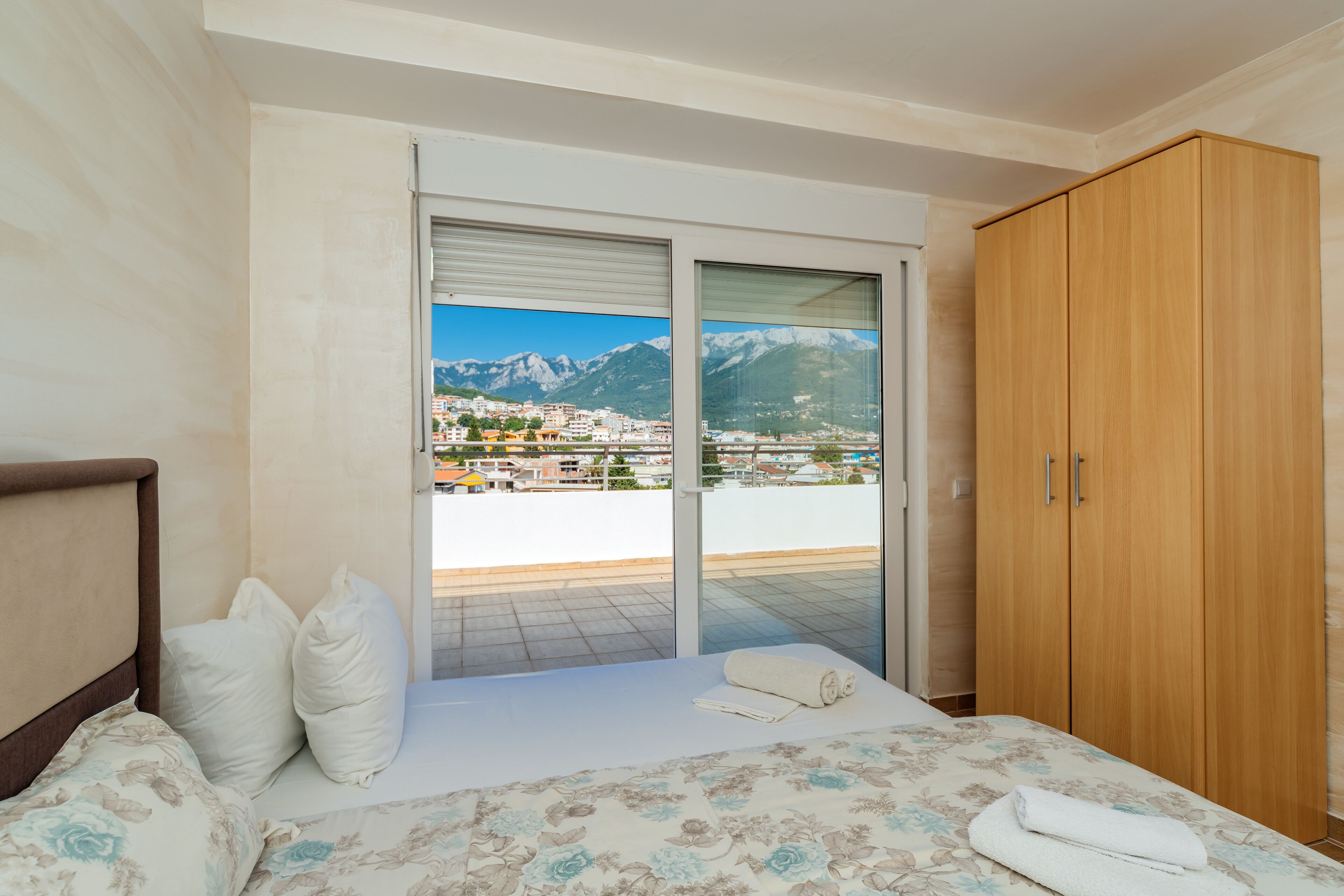 A bedroom with a bed, a wooden wardrobe, and a sliding glass door leading to a balcony with a mountain view.