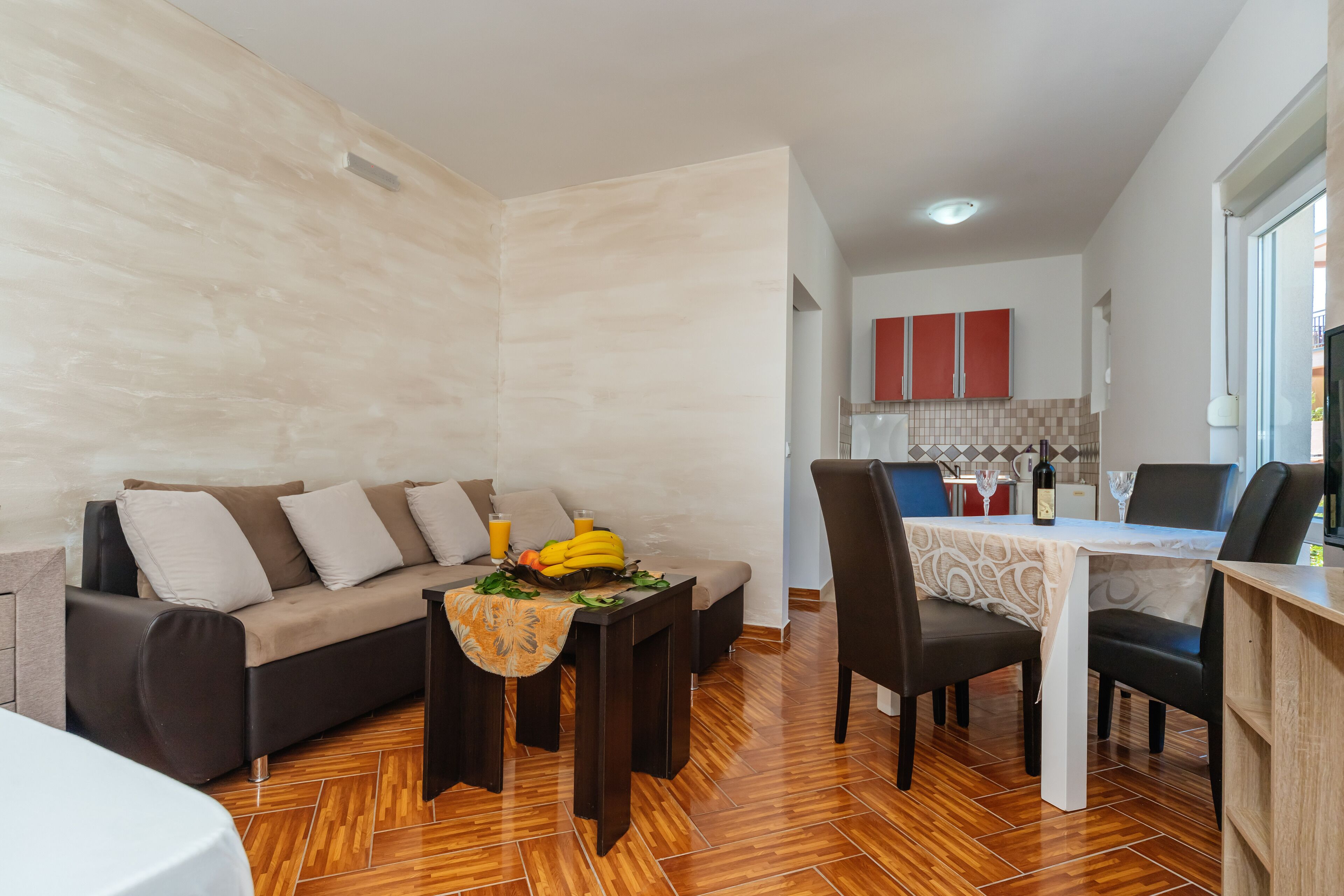 A modern living room with a sofa, dining table, and chairs. There is a kitchen area in the background with cabinets and a window.