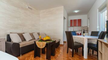 Una sala moderna con sofá, mesa de comedor y sillas. Se observa una cocina al fondo con armarios y una ventana.