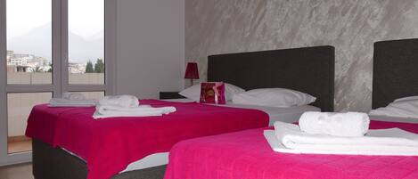 Habitación de hotel con dos camas, cada una con colcha rosa y toallas blancas. Se ve una ventana con vistas a edificios y una montaña.