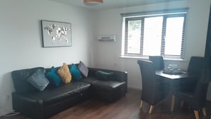 Un salon comprenant un canapé d’angle, une table à manger avec des chaises, une fenêtre avec des stores et un climatiseur fixé au mur.