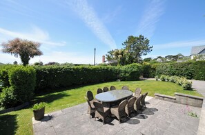 A patio with a round table and chairs, surrounded by a hedge and a grassy area.