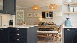 Une cuisine moderne avec des meubles de couleur sombre, une table à manger avec des chaises, des luminaires suspendus et une machine à café.