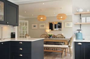 A modern kitchen with dark cabinetry, a dining table with chairs, pendant lights, and a coffee machine.